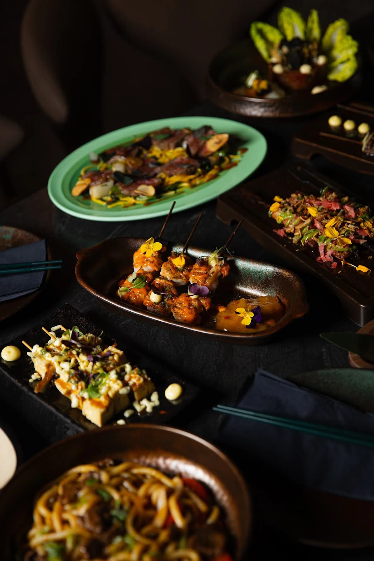 Een tafel met rijkgegarneerde Aziatische gerechten en spiesjes in warm, donker licht bij Anno Asia.