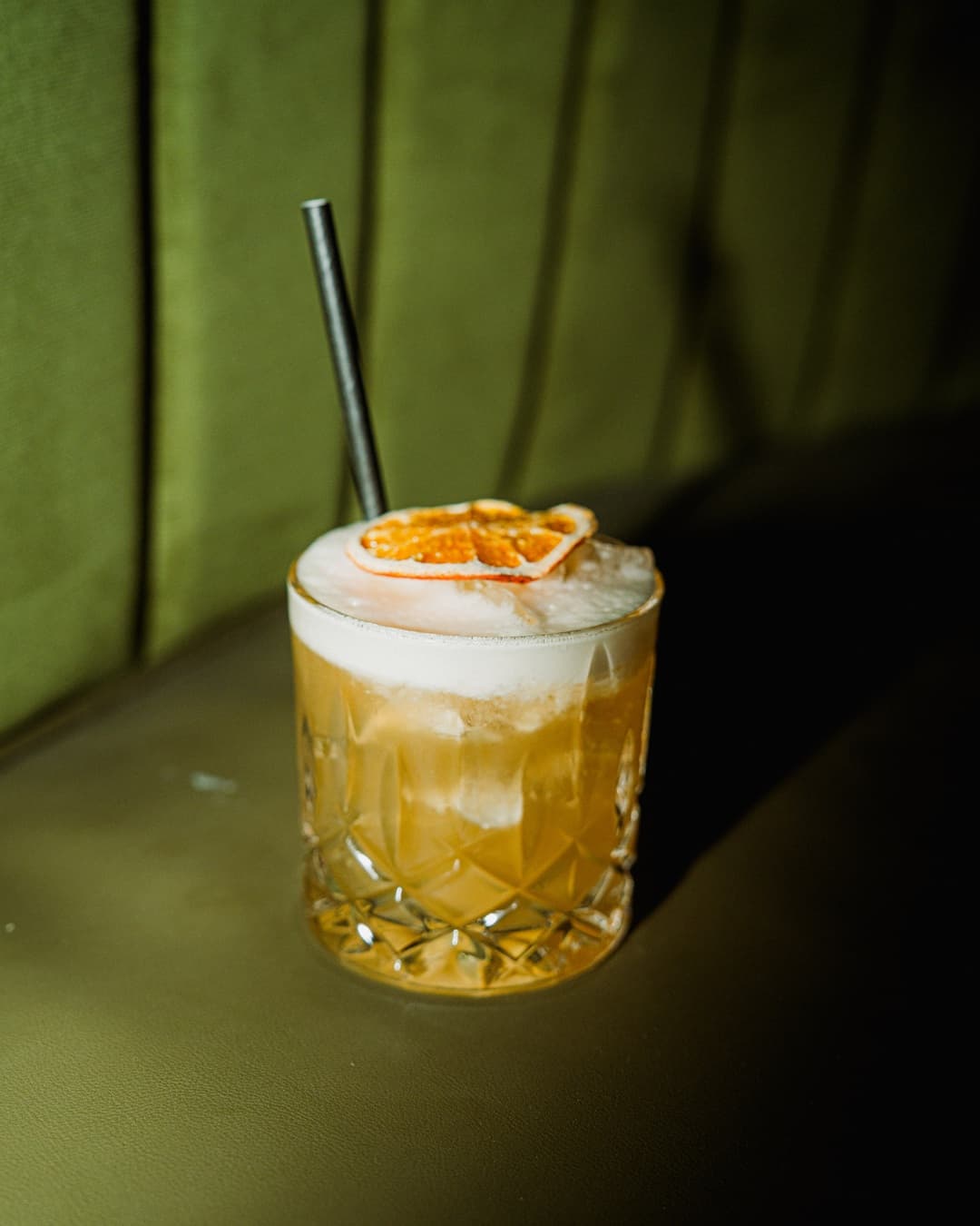 Een goudkleurige cocktail met schuim en gedroogde citrus tegen een groene bank bij Anno Asia.